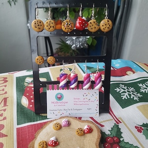 Puede incluir: Un conjunto de pendientes hechos a mano de arcilla polim&eacute;rica con varios dise&ntilde;os de comida, incluyendo galletas, tazas y bastones de caramelo. Los pendientes se muestran en un expositor de pendientes negro con una tarjeta blanca que dice "SGBoutique & Creations, Creative Fun & Eco-Elegance #DIY".