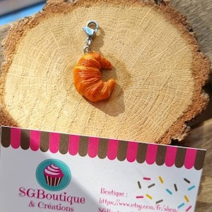 Puede incluir: Un charm que representa un peque&ntilde;o croissant naranja detallado con un lazo de metal plateado. El charm est&aacute; sobre una superficie de madera con una tarjeta de visita que dice "SG Boutique & Creations" e incluye un sitio web y una direcci&oacute;n de correo electr&oacute;nico.