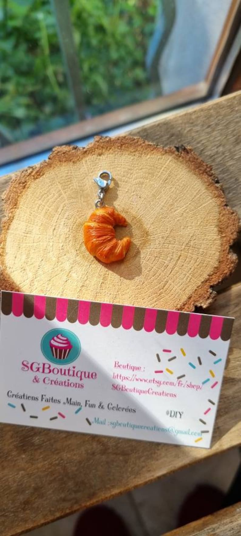 Puede incluir: Un charm que representa un peque&ntilde;o croissant naranja detallado con un lazo de metal plateado. El charm est&aacute; sobre una superficie de madera con una tarjeta de visita que dice "SG Boutique & Creations" e incluye un sitio web y una direcci&oacute;n de correo electr&oacute;nico.