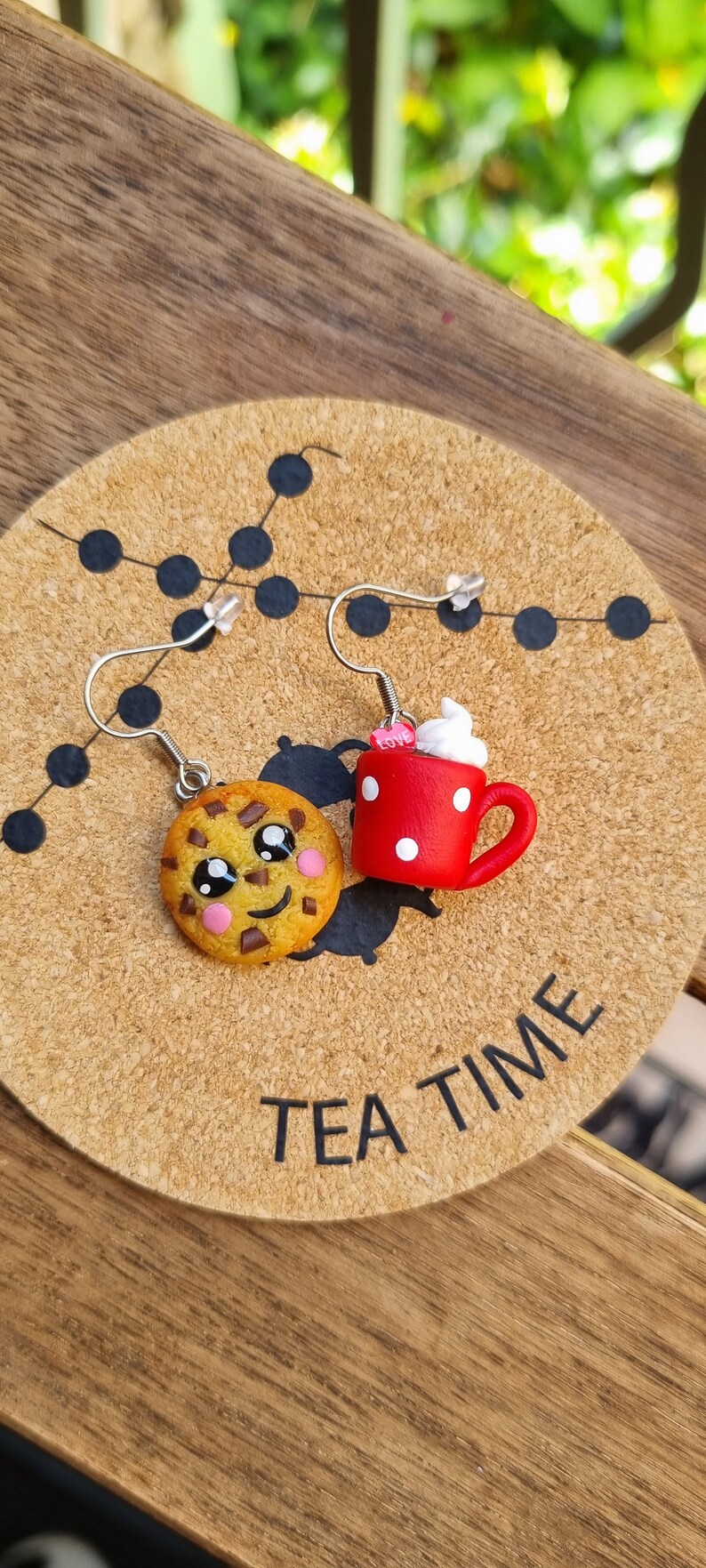 Puede incluir: Un par de pendientes con un dise&ntilde;o de taza roja y blanca con lunares, con un malvavisco blanco en la parte superior y una galleta de chocolate con chispas marr&oacute;n con una cara sonriente. Los pendientes se muestran en un posavasos de corcho con el texto "TEA TIME".
