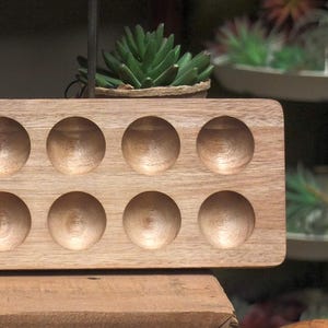 Puede incluir: Un portahuevos rectangular de madera clara con doce huecos circulares. El soporte tiene un acabado liso y natural. Está diseñado para sujetar los huevos de forma segura.