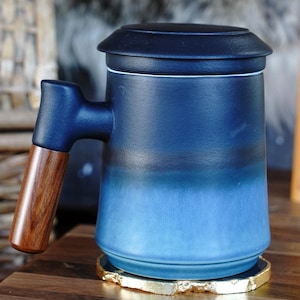 May include: A ceramic mug with a dark blue lid and handle, transitioning to a lighter blue base. The handle has a wooden grip. The mug sits on a gold-edged coaster. Books and a woven basket are in the background.