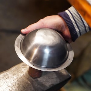 May include: A metal bowl with a circular rim is held by a person's hand. A wooden hammer is raised above the bowl, suggesting metalworking. The bowl is silver and has a smooth, reflective surface. The person is wearing a gray jacket with blue and white striped cuffs.