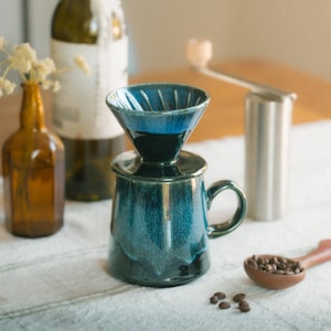 May include: A deep blue ceramic coffee dripper set resting on a matching mug. The glaze features a glossy finish with darker blue tones near the rim. Nearby are a brown glass bottle with dried flowers, a metal coffee grinder, and a wooden scoop.