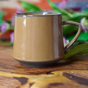 May include: A brown ceramic mug with a handle. The mug has a glossy finish and a dark brown base. The background includes a wooden surface and purple flowers. The mug is designed for hot beverages.