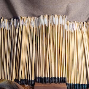 Puede incluir: Una gran colección de flechas de madera dispuestas en fila, cada una con astas de color claro, plumas blancas en los culatines y puntas de color oscuro. Las flechas se mantienen erguidas en una base de soporte de madera rectangular.