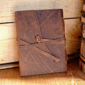 May include: A brown leather journal with a wrap-around strap closure. The journal is rectangular and appears to be handmade. The background includes wooden planks and a wooden barrel.