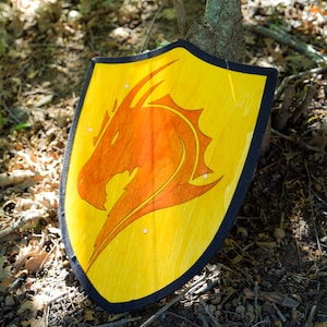 Könnte beinhalten: Ein gelber Schild mit schwarzem Rand und einer großen orangefarbenen Drachensilhouette in der Mitte. Der Schild hat eine spitze Unterseite und eine geschwungene Oberkante, mit kleinen weißen kreisförmigen Akzenten auf dem gelben Holz.