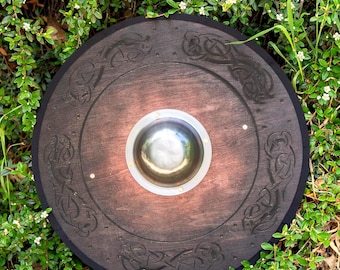 May include: A round wooden shield with a dark brown finish and intricate Celtic knotwork carvings around the edge. The shield features a central, silver-colored boss and is set against a backdrop of green foliage.