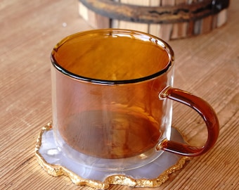 May include: A double-walled amber glass mug with a matching handle sits on a decorative coaster with a gold-colored edge. The mug is on a wooden surface, with a wooden barrel in the background.