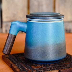 Puede incluir: Taza de cerámica azul degradado con asa y tapa de madera oscura. Se encuentra sobre un posavasos de madera tallada. La taza presenta un degradado de azul claro a oscuro.