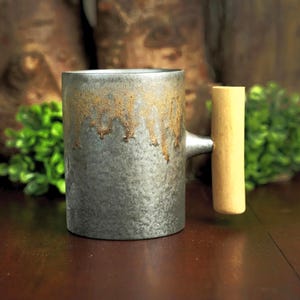 Könnte beinhalten: Ein zylindrischer Keramikbecher mit einer texturierten, metallisch grauen Oberfläche und einem dekorativen Tropfmuster in einem warmen Goldbraun am Rand. Der Becher verfügt über einen markanten, vertikalen, hellen Holzgriff an der Seite.