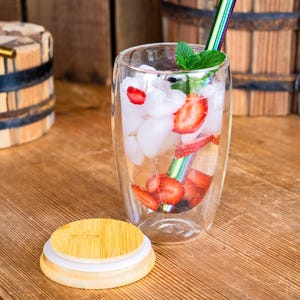 Puede incluir: Un vaso transparente de doble pared lleno de hielo, fresas en rodajas y menta. Una pajita arcoíris está en el vaso. Una tapa de madera está delante del vaso. La bebida está sobre una superficie de madera.