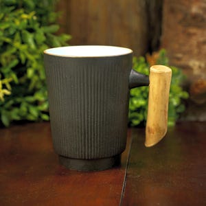 May include: A cylindrical mug with a matte black exterior featuring vertical ribbed texture and a smooth white interior. The mug has a distinct, light-colored, natural-looking wooden handle attached to the side, contrasting with the dark body.