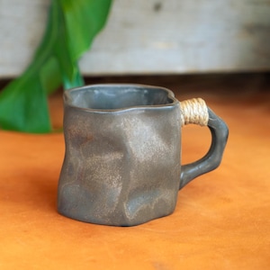 Könnte beinhalten: Dunkelgraue Keramiktasse mit strukturierter Oberfläche und einem mit Bindfaden umwickelten Henkel. Die Tasse hat eine ungewöhnliche, leicht unregelmäßige Form. Sie steht vor einem orangefarbenen Hintergrund, mit grünen Blättern im Hintergrund.