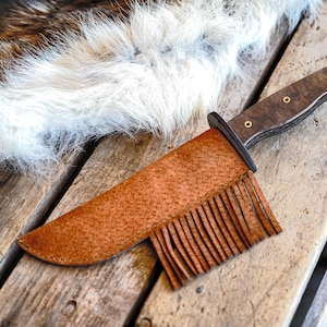 May include: A handcrafted hunting knife with a dark wooden handle and a brown leather sheath with fringe. The knife is resting on a weathered wooden surface, with a fur pelt in the background.