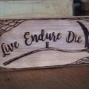 May include: Wooden sign featuring the words "Live Endure Die" and a scythe illustration. The text and image are burned into the wood, with a spiderweb design in the corners. The sign is rectangular and has a natural wood grain.