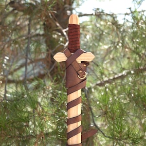 Könnte beinhalten: Ein Holzschwert mit einer Klinge und einem Parier aus hellem Naturholz. Der Griff ist mit einem dunkelbraunen, strukturierten Material umwickelt, und die obere Klinge ist mit einem dunkelbraunen Lederriemen befestigt, der einen messingfarbenen Ring aufweist.