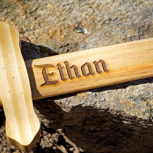 May include: A wooden toy sword with the name "Ethan" engraved on it. The sword is light brown and has a simple design.