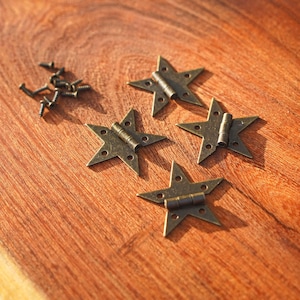 Puede incluir: Cuatro bisagras antiguas de bronce en forma de estrella con tornillos sobre una superficie de madera. Las bisagras están abiertas, revelando el mecanismo. La madera tiene un tono marrón rojizo cálido con veta visible.