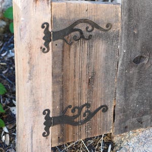 May include: Close-up of a rustic wooden gate with decorative black metal hinges. The hinges have a scrolled design, adding a touch of elegance to the weathered wood. The gate is made of vertical wooden planks.