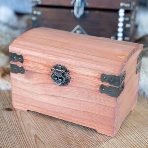 May include: A small, arched-top wooden chest with a reddish-brown finish. It features decorative metal hardware, including corner brackets and a clasp. Another chest is visible in the background.
