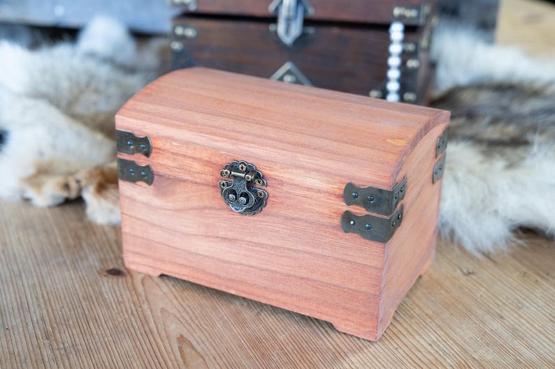 May include: A small, arched-top wooden chest with a reddish-brown finish. It features decorative metal hardware, including corner brackets and a clasp. Another chest is visible in the background.