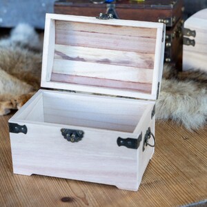 May include: An open, light-colored wooden chest with a hinged lid. The chest has metal hardware and a handle. The interior of the lid is made of wooden slats. The chest is sitting on a wooden surface.