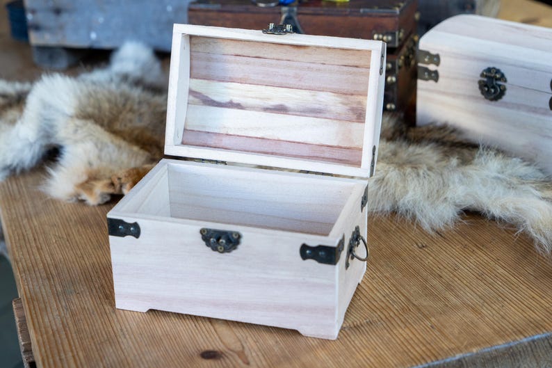 May include: An open, light-colored wooden chest with a hinged lid. The chest has metal hardware and a handle. The interior of the lid is made of wooden slats. The chest is sitting on a wooden surface.