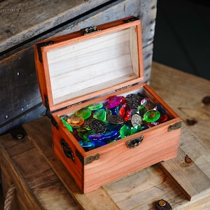 May include: A wooden treasure chest with a hinged lid, filled with colorful glass gems and antique-style coins. The chest is made of reddish-brown wood with metal corner protectors and a clasp. The gems are various colors, including green, blue, red, and purple.