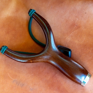 Puede incluir: Un tirachinas de madera de color marrón oscuro pulido con un marco en forma de Y y un mango ergonómico liso. Incluye una banda elástica de color verde oscuro unida a las dos puntas superiores y una pequeña bolsa de cuero negro.