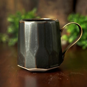 May include: A dark charcoal gray ceramic mug with a faceted, geometric exterior surface. The mug features a metallic gold-toned handle and a contrasting white base. The rim and edges of the facets show subtle brown highlights against the dark finish.