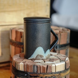 May include: A black ceramic mug with a lid and handle. The mug has a mountain design in light blue and brown at the base. The mug is approximately 13 cm tall and rests on a wooden surface.