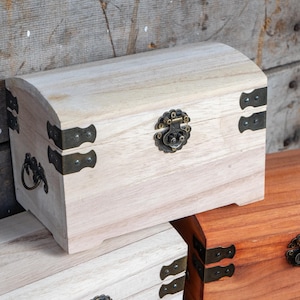 May include: A light-colored wooden chest with a curved top, metal hinges, and a decorative clasp. The chest is part of a set, with another chest visible in the background. The chests are made of wood and have metal hardware.