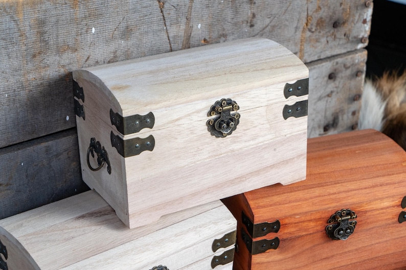May include: A light-colored wooden chest with a curved top, metal hinges, and a decorative clasp. The chest is part of a set, with another chest visible in the background. The chests are made of wood and have metal hardware.