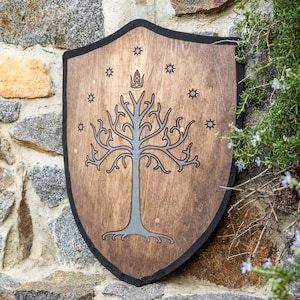 Pode incluir: Um escudo de madeira com acabamento castanho escuro e borda preta. O centro apresenta um gráfico de árvore estilizado em cinza encimado por uma coroa, rodeado por sete pequenos símbolos de estrelas dispostos em arco no topo.