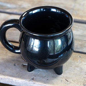 May include: A black ceramic cauldron mug with a handle. The mug has three small feet and is sitting on a wooden surface.