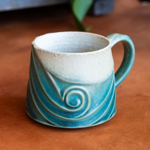 Peut inclure: Une tasse en céramique faite à la main avec un motif de vagues turquoise et un dessus blanc. La tasse a une anse incurvée et mesure environ 10 cm de haut.
