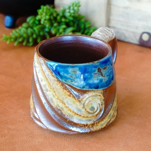 May include: Handmade ceramic mug with a brown, blue, and tan swirl design. The mug features a blue band with a bird design and a twisted handle. The interior is dark brown.