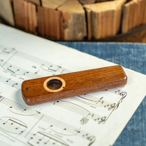 May include: A wooden kazoo with a circular sound hole and a light-colored wood accent. The kazoo rests on sheet music and a blue denim surface. A wooden basket is in the background.