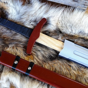 May include: A wooden sword with a red handle and a black grip, resting on a faux fur surface. The sword is partially sheathed in a silver scabbard, and a red scabbard with black straps lies below.