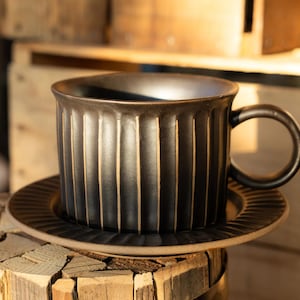 May include: A dark charcoal ceramic mug and matching saucer set featuring a vertical ribbed texture. The mug has a rounded handle and a wide rim, while the saucer displays a radial grooved pattern. Both pieces show light brown edges.