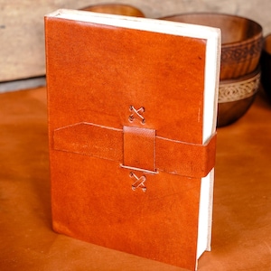 May include: A handmade leather journal with a rich, orange-brown hue. The journal features a wraparound strap closure secured with decorative cross-stitch details. The pages are visible, and the journal is set against a warm, textured background.