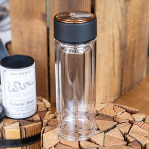 May include: A clear glass tea infuser bottle with a black lid and a gold rim. The bottle is sitting on a wooden surface.
