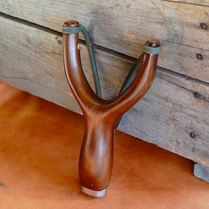 May include: A handcrafted wooden slingshot with a polished, dark brown finish. The Y-shaped frame features a smooth, curved design, with green rubber bands and a silver metal base. The slingshot is set against a rustic wooden background.