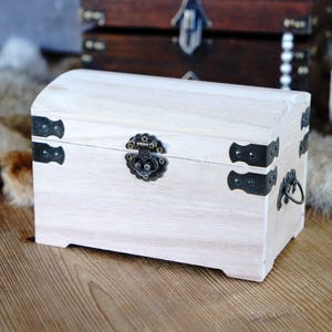 May include: A small, light-colored wooden chest with a curved top. The chest features black metal hardware, including corner brackets, a clasp, and a handle. Other similar chests are visible in the background.