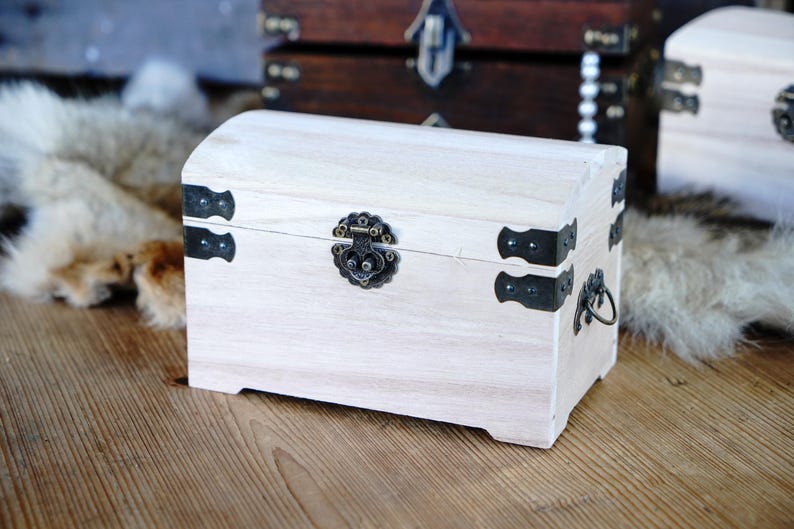 May include: A small, light-colored wooden chest with a curved top. The chest features black metal hardware, including corner brackets, a clasp, and a handle. Other similar chests are visible in the background.