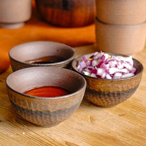 Puede incluir: Tres cuencos pequeños de cerámica marrón, dos llenos de un líquido rojo oscuro y uno lleno de cebolla roja picada.