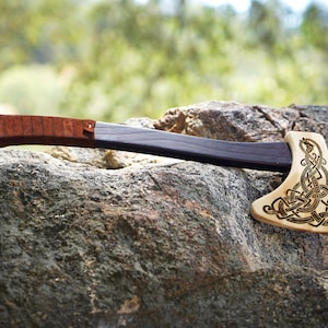 May include: A hand axe with a light-colored wooden head featuring an intricate black engraved knotwork pattern. The handle is made of dark wood and is partially wrapped in a brown leather cord near the end, providing a textured grip.