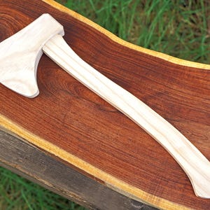 May include: A light-colored wooden axe with a curved handle and a wide blade. The axe is resting on a dark brown wooden surface. The axe head is rectangular with a curved cutting edge.